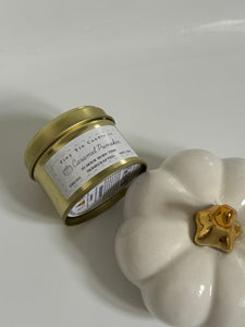 Tiny Tin Candle Co.'s Caramel Pumpkin soy wax candle in a gold tin, displayed next to a decorative white pumpkin with a golden stem. The candle, featuring a 23-hour burn time, is vegan and handcrafted, offering a warm autumn scent to complement the cosy, seasonal décor