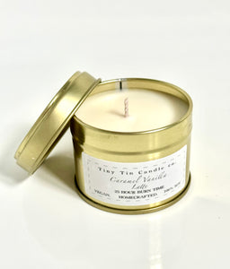 Tiny Tin Candle Co.'s Caramel Vanilla Latte soy wax candle with an open tin lid. The handcrafted, vegan candle sits on a smooth surface, showcasing its clean and minimalistic gold tin packaging with a white label. The candle promises 23 hours of burn time, offering a warm and cozy caramel vanilla scent