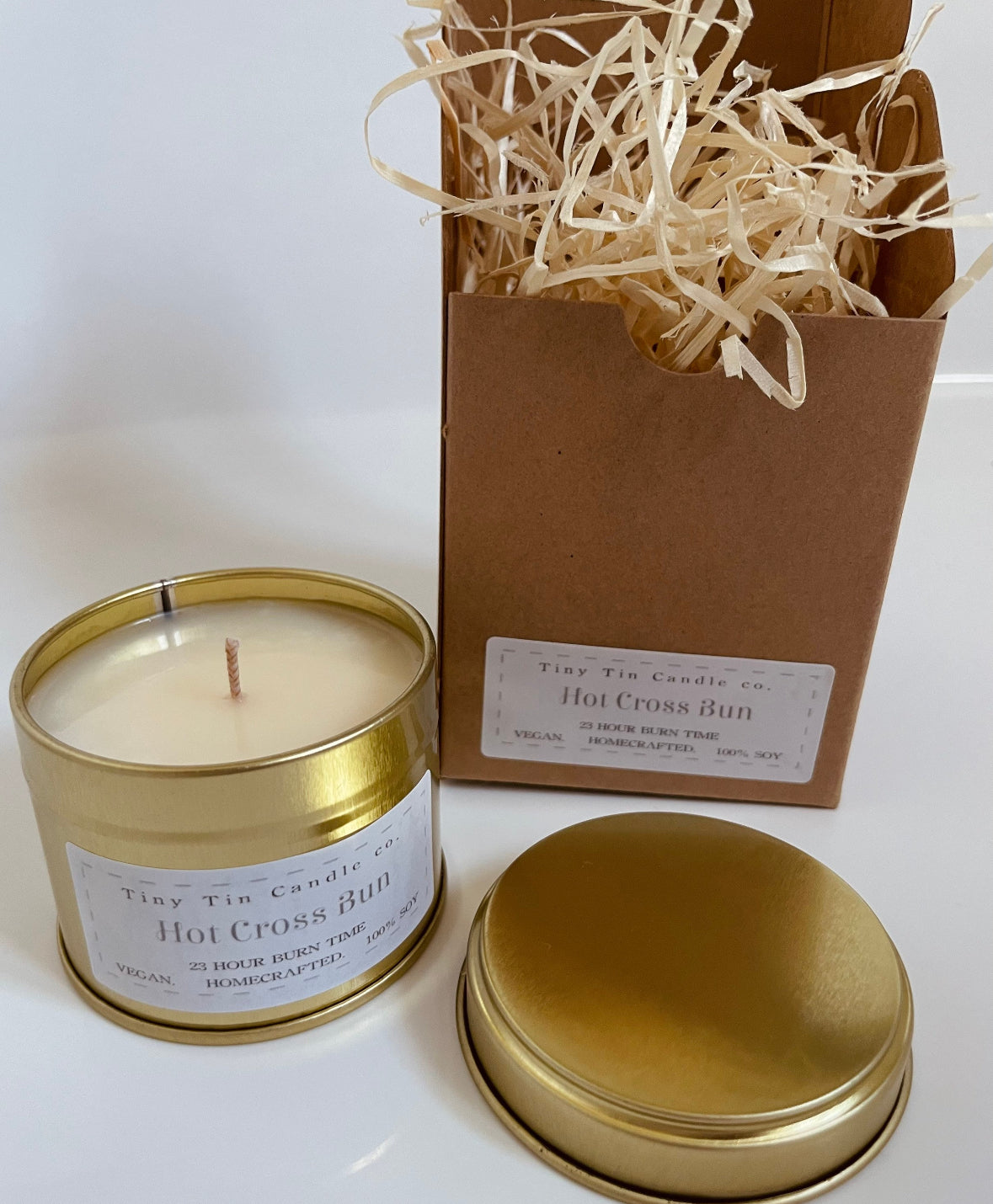 A handmade 'Hot Cross Bun' scented candle by Tiny Tin Candle Co., elegantly packaged in a tin with a Kraft paper box filled with wood shavings, epitomising artisanal charm