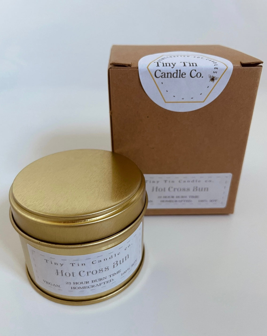 A handmade 'Hot Cross Bun' scented candle by Tiny Tin Candle Co., elegantly packaged in a tin with a Kraft paper box filled with wood shavings, epitomising artisanal charm