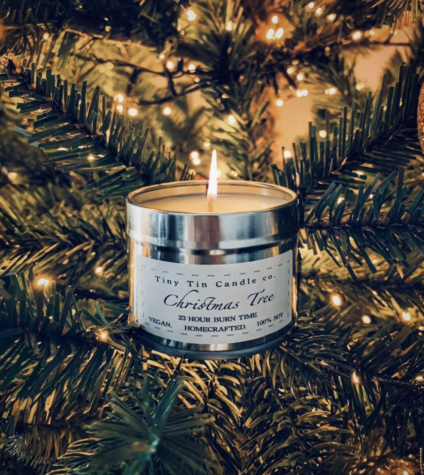 A lit homemade Christmas Tree scented candle from Tiny Tin Candle Co., nestled among the lush branches of a decorated Christmas tree. The candle's silver tin glimmers with the reflection of twinkling tree lights, highlighting the label which promises a 23-hour burn time and boasts its homecrafted, 100% soy quality, adding a festive and artisanal touch to the holiday decor