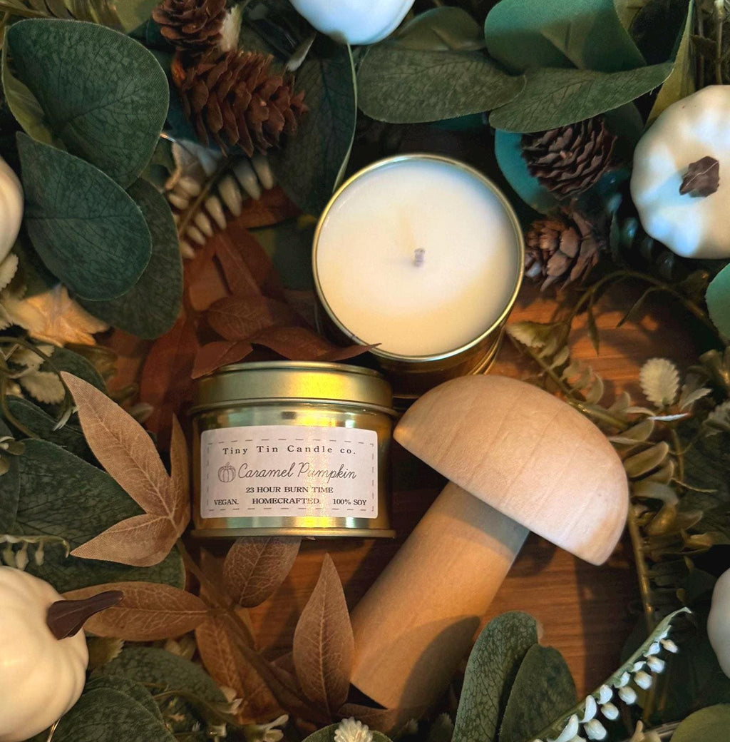 An autumn-inspired scene featuring Tiny Tin Candle Co.'s Caramel Pumpkin soy wax candle. The candle is presented in a gold tin with its lid off, surrounded by decorative pumpkins and vibrant fall leaves. The setup showcases the cosy, handcrafted candle, which promises a 23-hour burn time and is vegan and 100% soy-based, perfect for enhancing the seasonal ambiance with a warm caramel pumpkin scent
