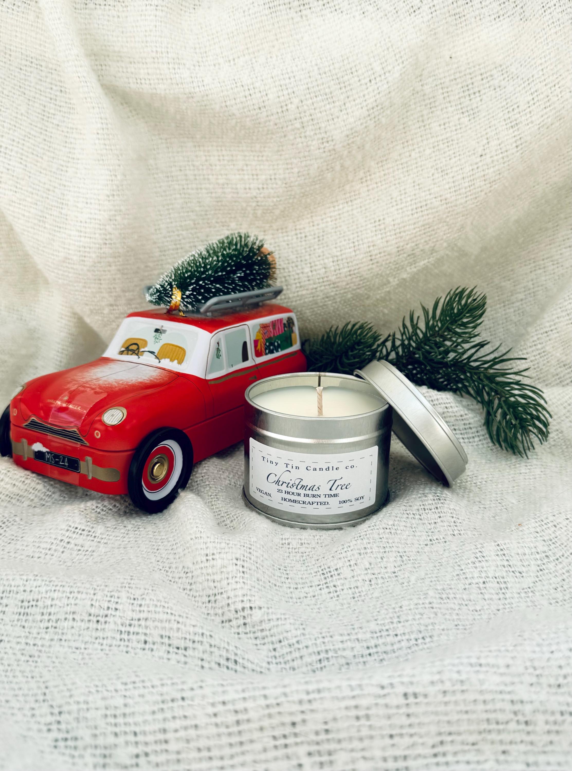 A lit homemade Christmas Tree scented candle from Tiny Tin Candle Co., nestled among the lush branches of a decorated Christmas tree. The candle's silver tin glimmers with the reflection of twinkling tree lights, highlighting the label which promises a 23-hour burn time and boasts its homecrafted, 100% soy quality, adding a festive and artisanal touch to the holiday decor