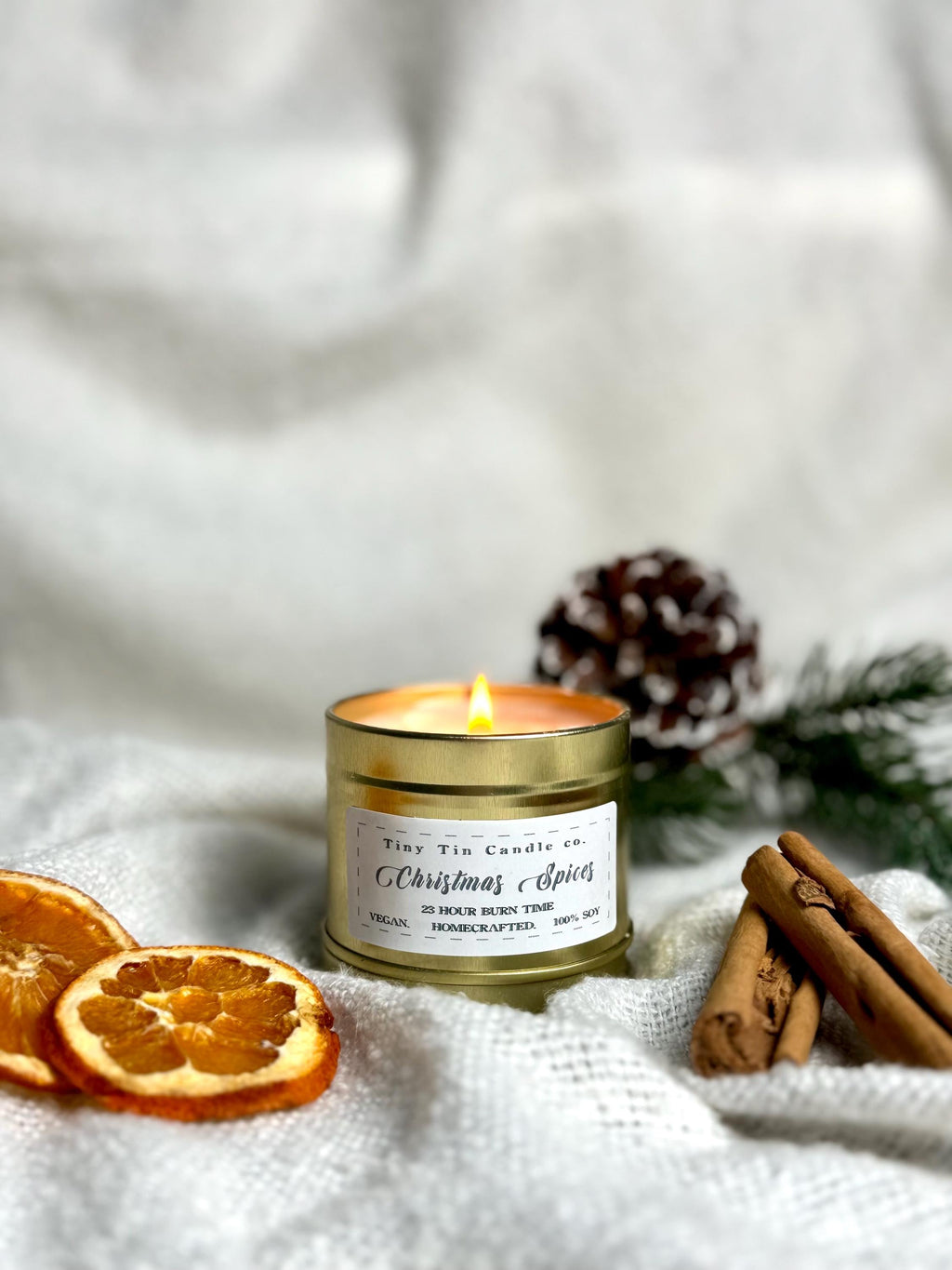 The Christmas Spices homemade candle from Tiny Tin Candle Co. alight in a golden tin, surrounded by cinnamon sticks, dried oranges, and pine, evoking a rich, artisanal holiday scent