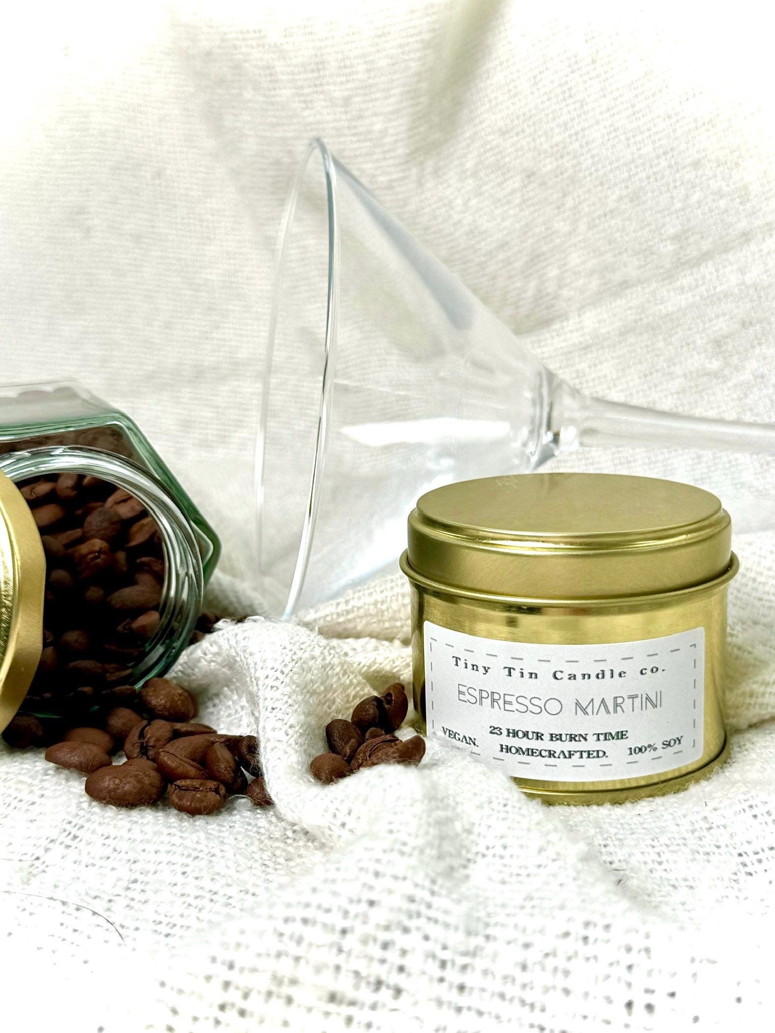 Handmade Espresso Martini scented soy candle from Tiny Tin Candle Co., featuring coffee beans on top, with a 23-hour burn time label, vegan and homecrafted, in a stacked gold tin against a backdrop of lush greenery