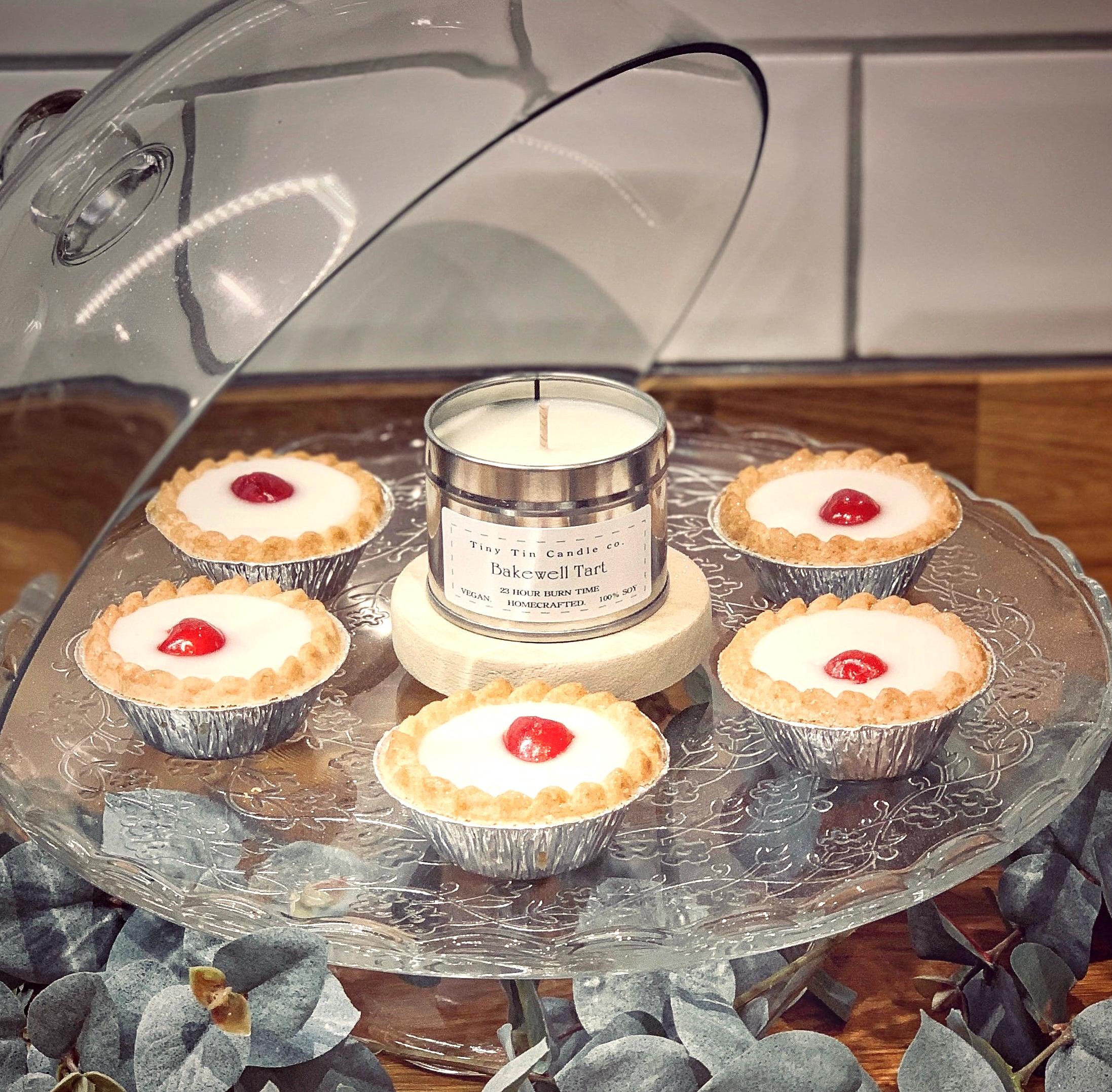 Tiny Tin Candle Co.'s 'Bakewell Tart' homemade candle is elegantly presented on a cake stand surrounded by actual Bakewell tarts, blending the art of home baking with the craft of candle-making.