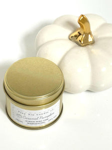 Tiny Tin Candle Co.'s Caramel Pumpkin soy wax candle in a gold tin, displayed next to a decorative white pumpkin with a golden stem. The candle, featuring a 23-hour burn time, is vegan and handcrafted, offering a warm autumn scent to complement the cosy, seasonal d√©cor