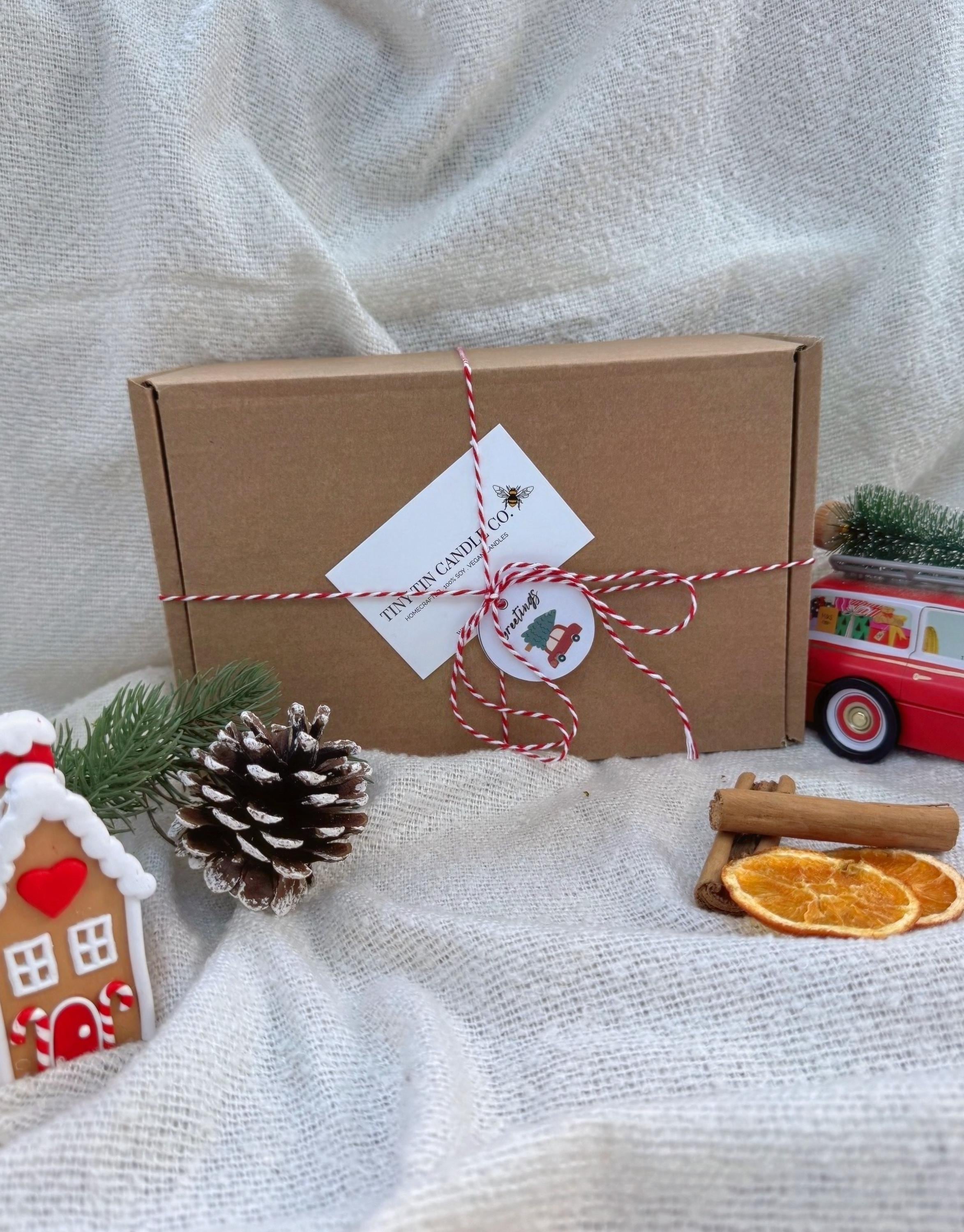 The image showcases a beautifully lit "Christmas Spices" candle from Tiny Tin Candle Co., surrounded by festive elements. The candle is nestled among natural holiday decorations, including cinnamon sticks, dried orange slices, small pumpkins, and sprigs of pine greenery. The warm glow from the candle enhances the cozy and inviting atmosphere, perfect for setting the mood during the Christmas season. The label emphasizes that the candle is 100% soy, vegan, handcrafted, and offers a 23-hour burn time.