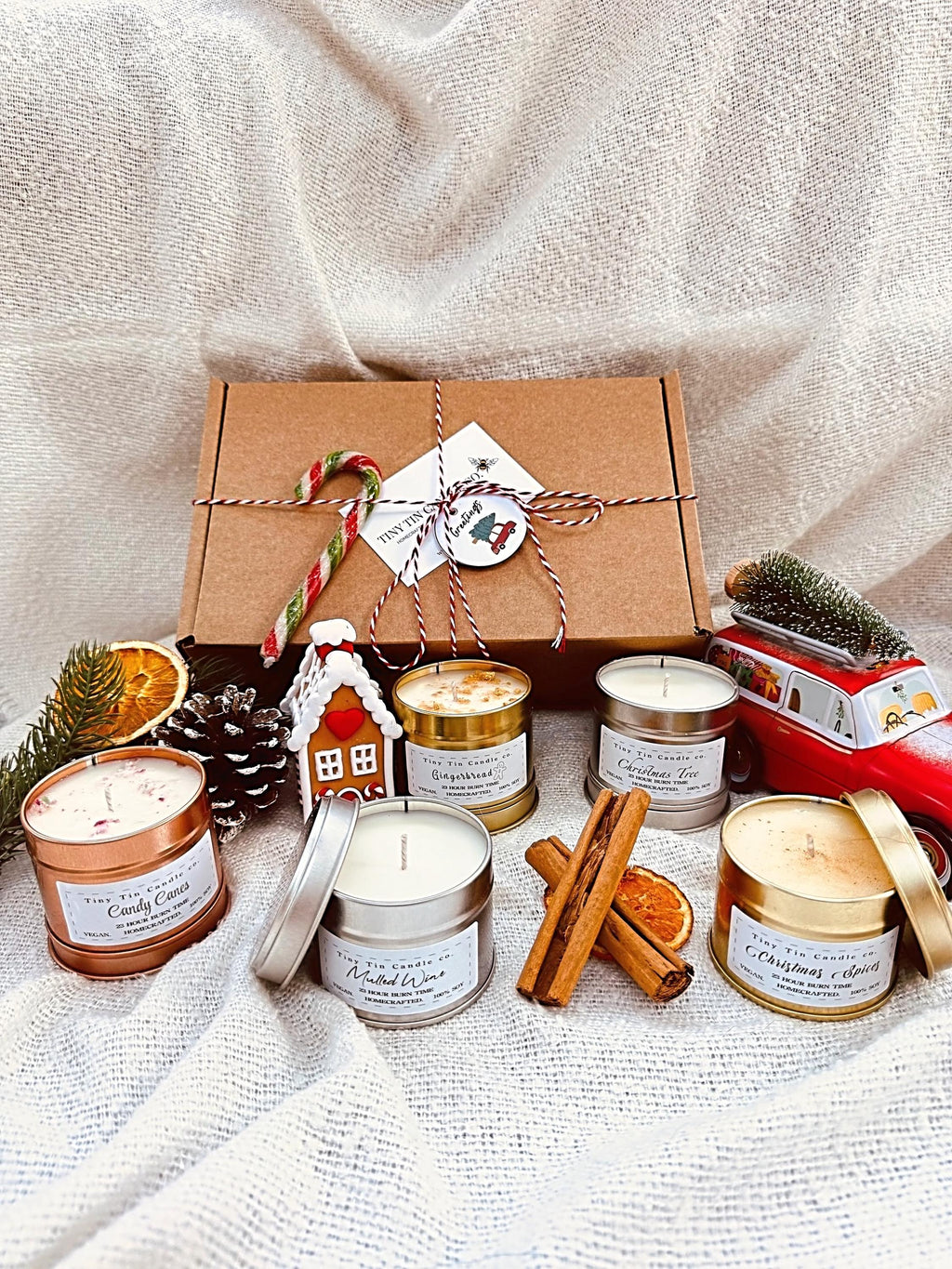 The image features a beautifully arranged gift box from Tiny Tin Candle Co., showcasing five festive candles nestled in shredded paper. The candles include "Christmas Spices," "Christmas Tree," "Candy Canes," "Gingerbread," and "Mulled Wine." Each candle is labeled with its burn time of 23 hours and highlights that they are 100% soy, vegan, and handcrafted. The arrangement is completed with a Tiny Tin Candle Co. business card and is framed with frosted greenery, making it a perfect holiday gift set to bring