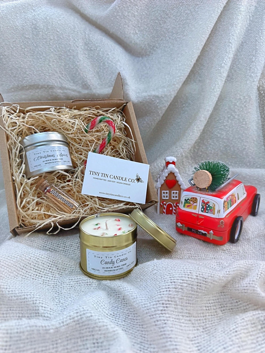 The image shows a beautifully presented Tiny Tin Candle Co. gift box. Inside the box are two small tin candles labeled "Christmas Spices" and "Candy Cane," nestled in shredded paper. The display is decorated with festive touches, including dried orange slices, a small orange pumpkin, candy canes, a sprig of mistletoe, and a small glass bottle of matches. Soft fairy lights are woven around the box, adding a warm, cozy glow, perfect for the holiday season.