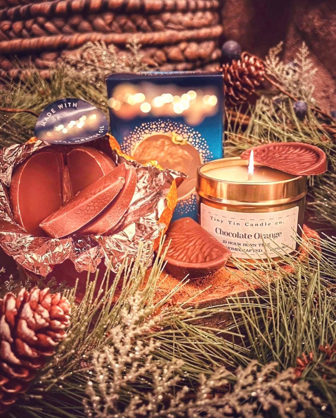 The Chocolate Orange homemade candle from Tiny Tin Candle Co. is lit, casting a cozy glow on a holiday setting with pine needles and cones. A chocolate orange piece sits nearby, complementing the candle's sweet aroma. Festive lights twinkle in the background, creating a warm, artisanal ambiance