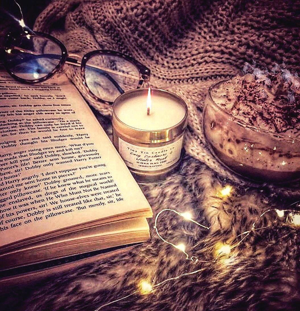 A warm, homemade candle flickers beside an open book and a pair of glasses, on a fluffy faux fur surface. A cozy blanket and a cup of whipped hot chocolate add to the inviting, relaxed setting