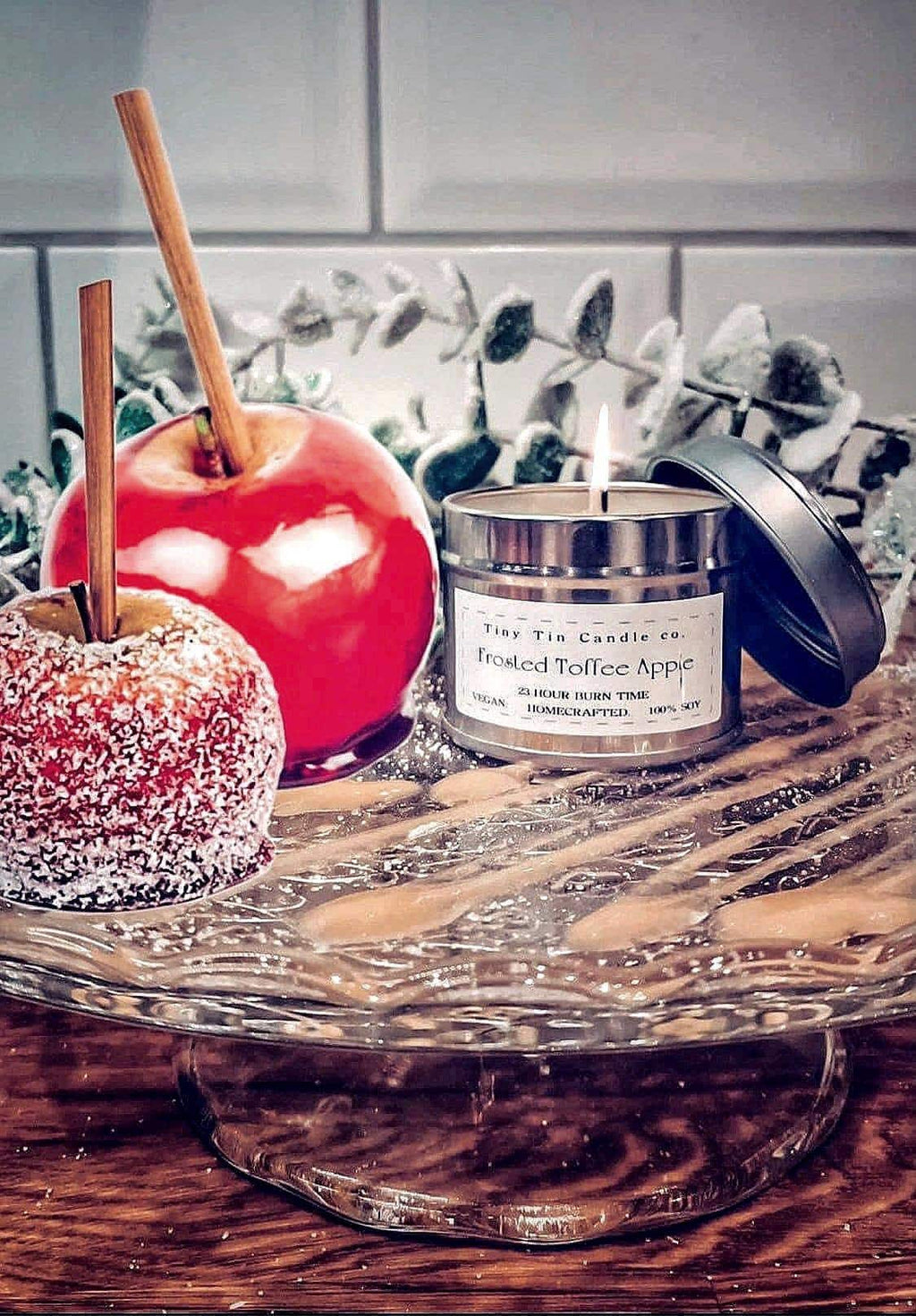 An inviting scene featuring a lit homemade candle by Tiny Tin Candle Co., labeled 'Frosted Toffee Apple' with a 23-hour burn time, alongside a festive candy apple and a sugar-frosted toffee apple. The candle, crafted from 100% soy, sits elegantly on a wooden surface, its open silver tin reflecting the cozy ambience, surrounded by a dusting of frosty decorations, capturing the essence of a warm, sweet treat on a cool day.