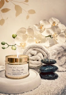 iny Tin Candle Co.'s Spa Day homemade scented candle, elegantly placed beside spa stones, orchids, and fluffy towels, evoking a serene, indulgent atmosphere.