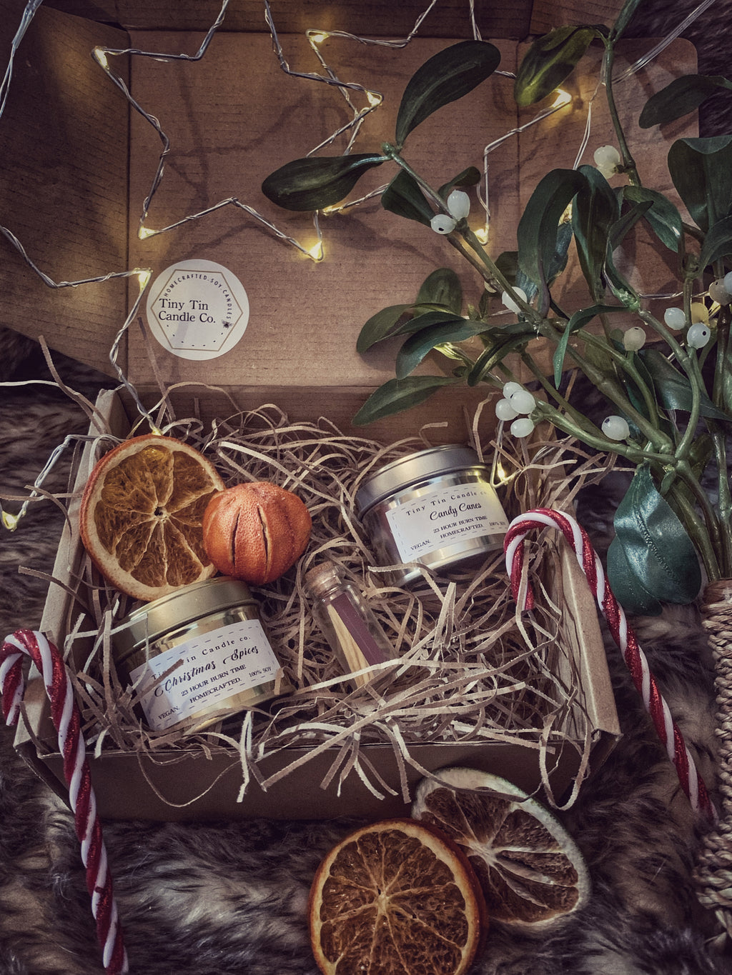 The image shows a beautifully presented Tiny Tin Candle Co. gift box. Inside the box are two small tin candles labeled "Christmas Spices" and "Candy Cane," nestled in shredded paper. The display is decorated with festive touches, including dried orange slices, a small orange pumpkin, candy canes, a sprig of mistletoe, and a small glass bottle of matches. Soft fairy lights are woven around the box, adding a warm, cozy glow, perfect for the holiday season.