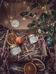 The image shows a beautifully presented Tiny Tin Candle Co. gift box. Inside the box are two small tin candles labeled "Christmas Spices" and "Candy Cane," nestled in shredded paper. The display is decorated with festive touches, including dried orange slices, a small orange pumpkin, candy canes, a sprig of mistletoe, and a small glass bottle of matches. Soft fairy lights are woven around the box, adding a warm, cozy glow, perfect for the holiday season.