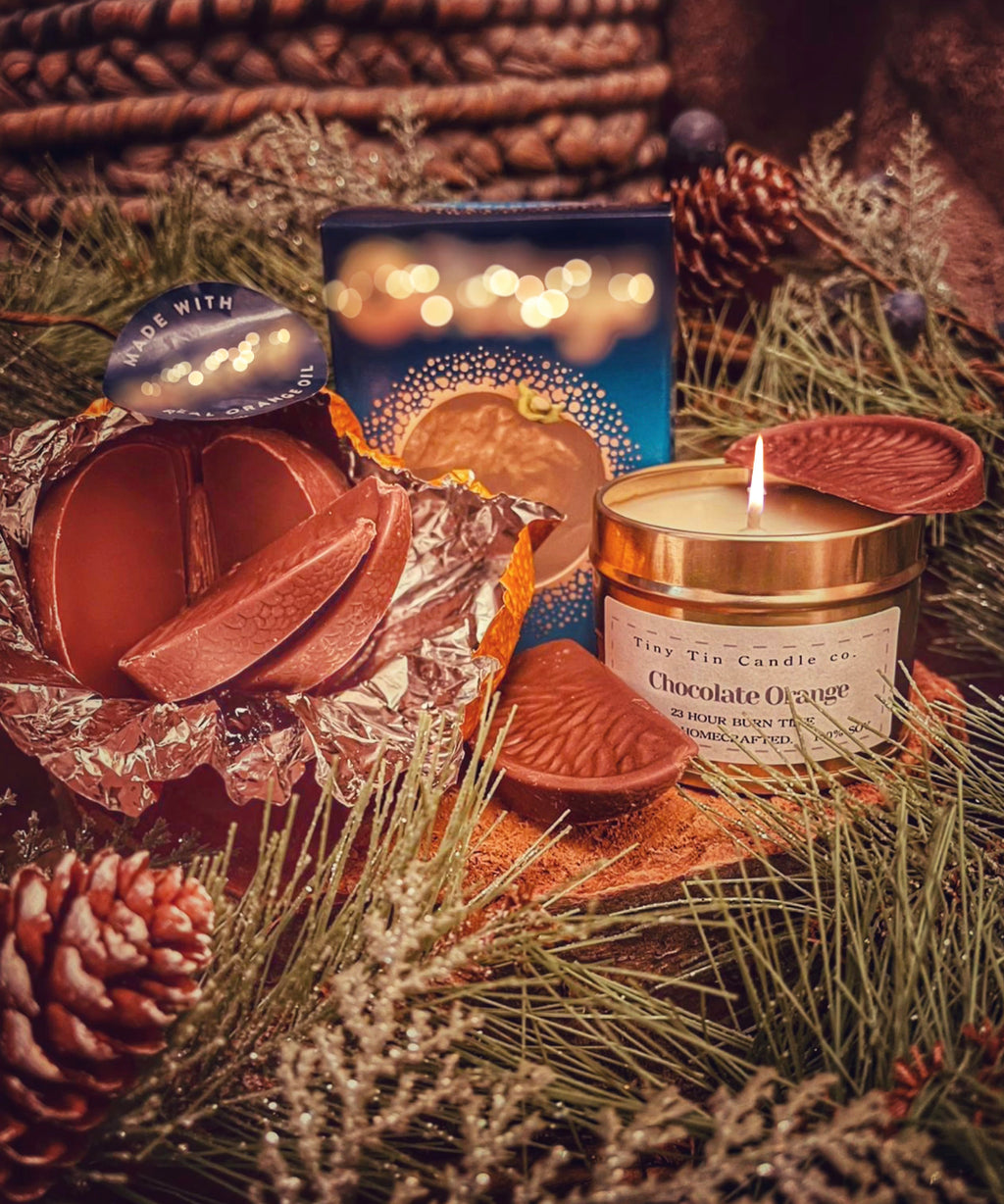 The Chocolate Orange homemade candle from Tiny Tin Candle Co. is lit, casting a cozy glow on a holiday setting with pine needles and cones. A chocolate orange piece sits nearby, complementing the candle's sweet aroma. Festive lights twinkle in the background, creating a warm, artisanal ambiance