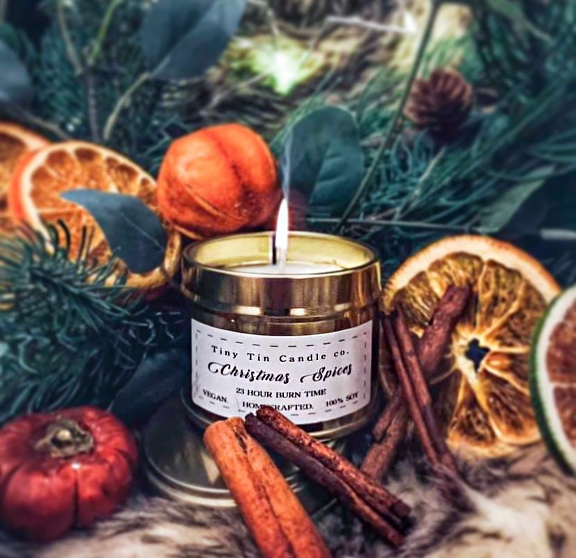 The image showcases a beautifully lit "Christmas Spices" candle from Tiny Tin Candle Co., surrounded by festive elements. The candle is nestled among natural holiday decorations, including cinnamon sticks, dried orange slices, small pumpkins, and sprigs of pine greenery. The warm glow from the candle enhances the cozy and inviting atmosphere, perfect for setting the mood during the Christmas season. The label emphasizes that the candle is 100% soy, vegan, handcrafted, and offers a 23-hour burn time.