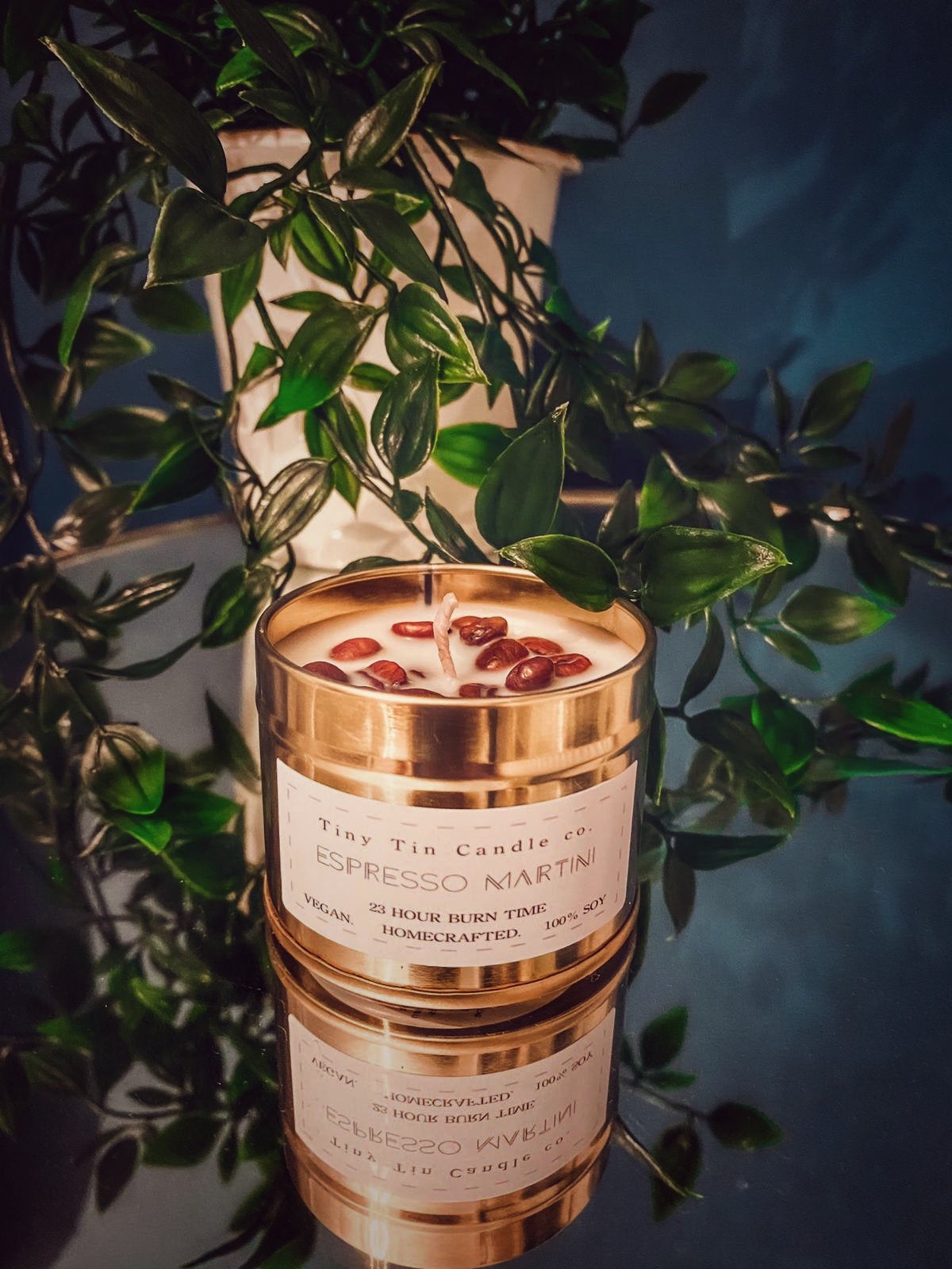 Handmade Espresso Martini scented soy candle from Tiny Tin Candle Co., featuring coffee beans on top, with a 23-hour burn time label, vegan and homecrafted, in a stacked gold tin against a backdrop of lush greenery