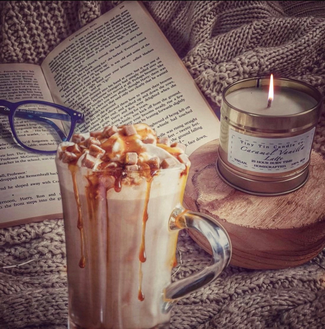 Cosy autumn scene featuring Tiny Tin Candle Co.'s hand-poured soy wax candle in Caramel Vanilla Latte scent. The candle, placed on a wooden coaster, burns softly, surrounded by a knit blanket, an open book, and a warm caramel-topped latte. Perfect for creating a comforting, sweet, and inviting atmosphere