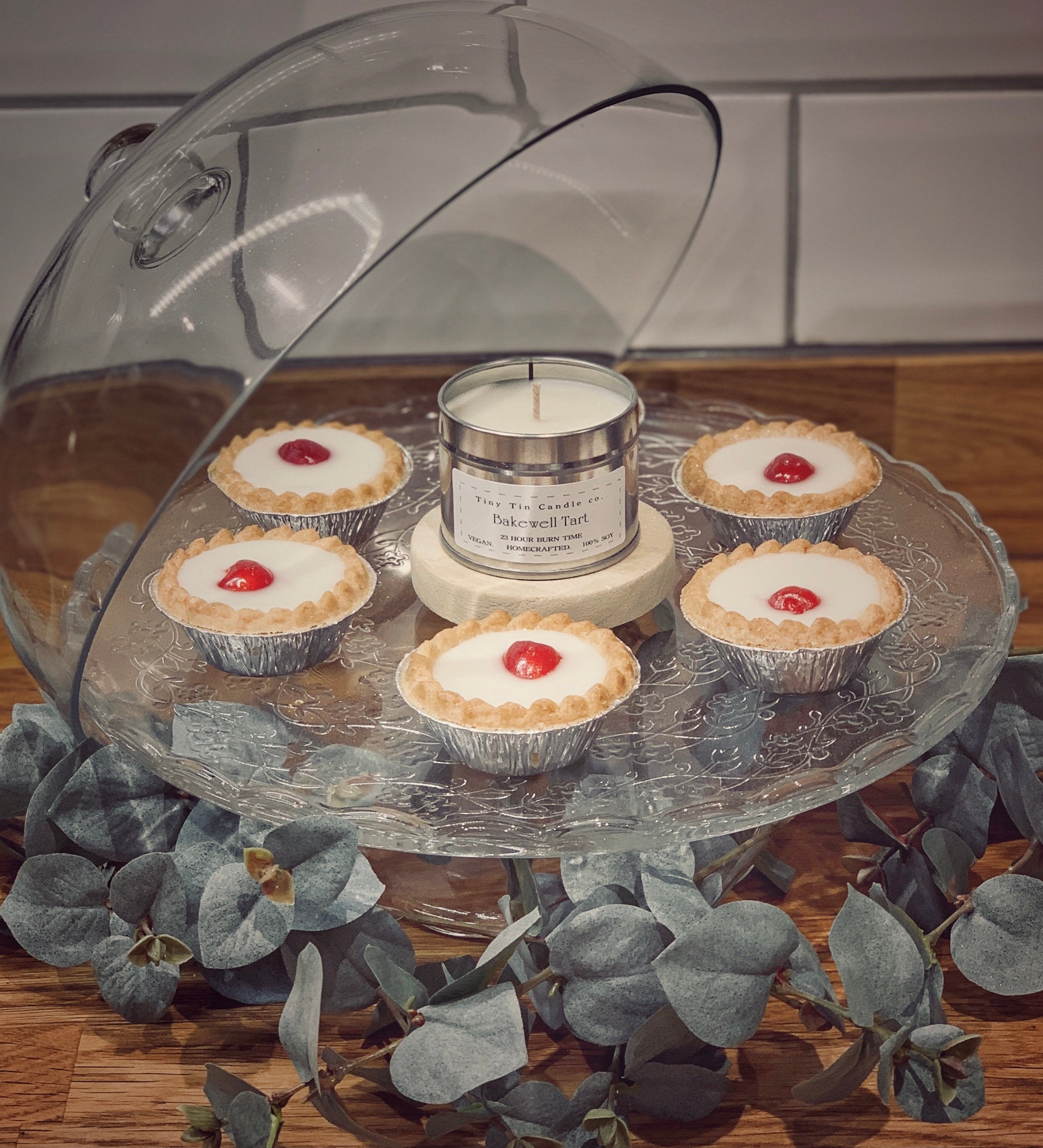 Tiny Tin Candle Co.'s 'Bakewell Tart' homemade candle is elegantly presented on a cake stand surrounded by actual Bakewell tarts, blending the art of home baking with the craft of candle-making.