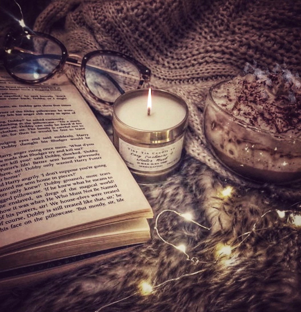 A warm, homemade candle flickers beside an open book and a pair of glasses, on a fluffy faux fur surface. A cozy blanket and a cup of whipped hot chocolate add to the inviting, relaxed setting