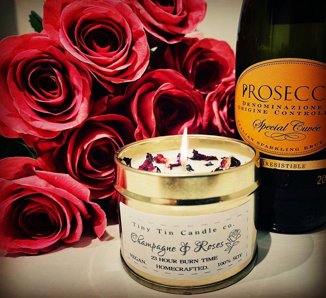 Celebrate with Tiny Tin Candle Co.'s 'Champagne & Roses' homemade soy candle, lit beside a bouquet of red roses and a bottle of Prosecco, setting a romantic and festive mood