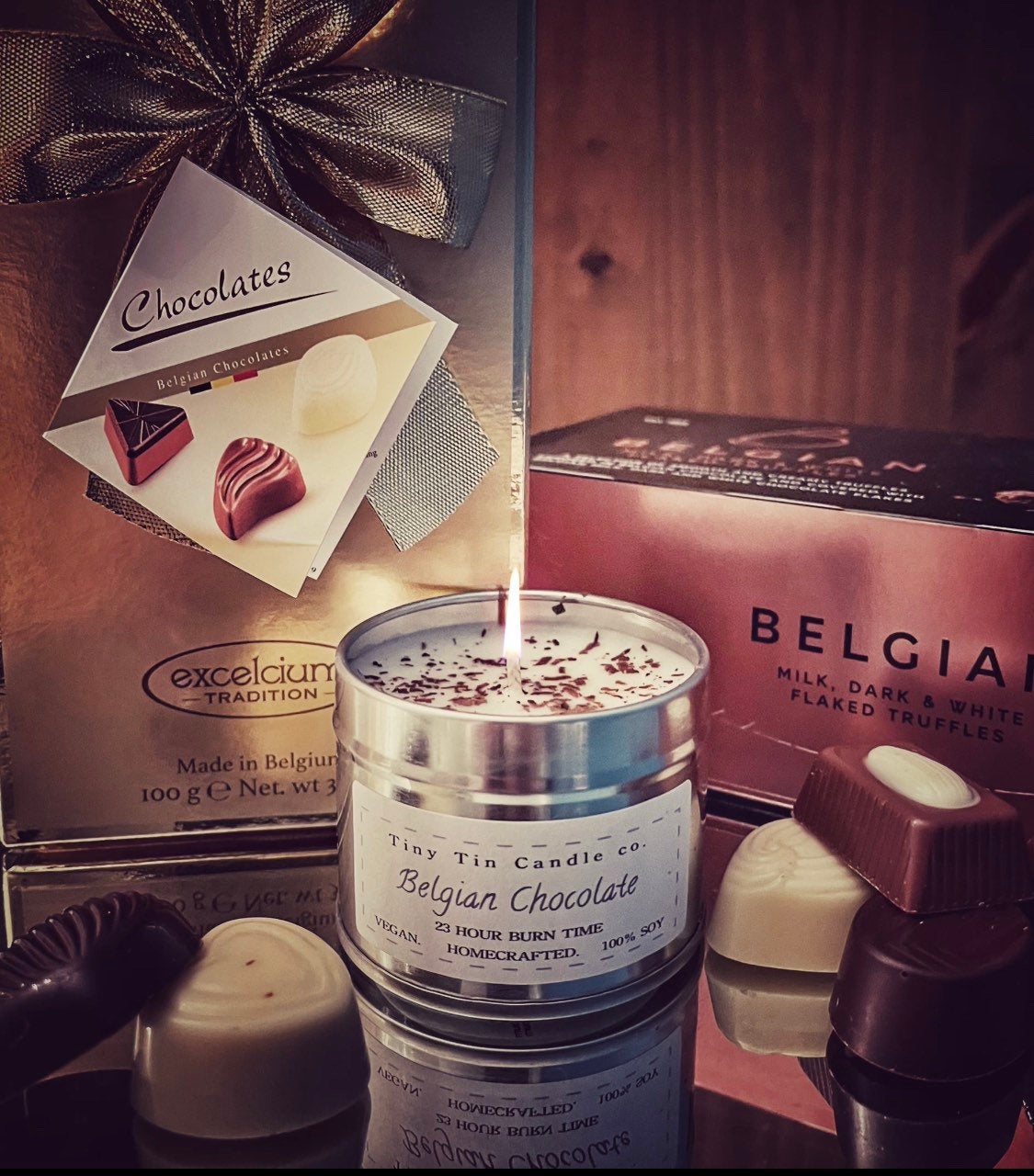 A lit 'Belgian Chocolate' homemade soy candle from Tiny Tin Candle Co. sits amidst an assortment of real Belgian chocolates, conveying a luxurious and richly scented experience