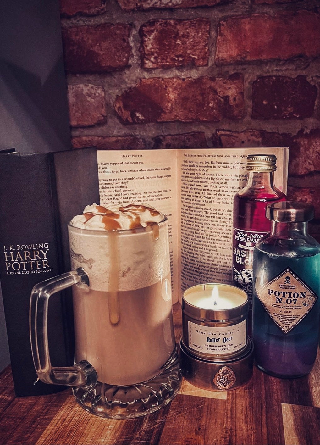 A cosy setting with Tiny Tin Candle Co.'s 'Butter Beer' handmade candle lit beside a frothy mug of butterbeer, a Harry Potter book, and whimsical potion bottles, evoking a magical ambiance