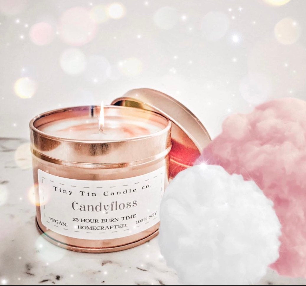 Tiny Tin Candle Co.'s Candyfloss homemade candle, with a lit wick casting a soft glow amidst a dreamy backdrop of bokeh lights. The label proudly states 'Vegan, 23 hour burn time, homecrafted, 100% soy', embodying the luxurious and artisanal essence of the candle, accompanied by fluffy white and pink candy floss clouds, evoking the sweet, sugary scent of the candle