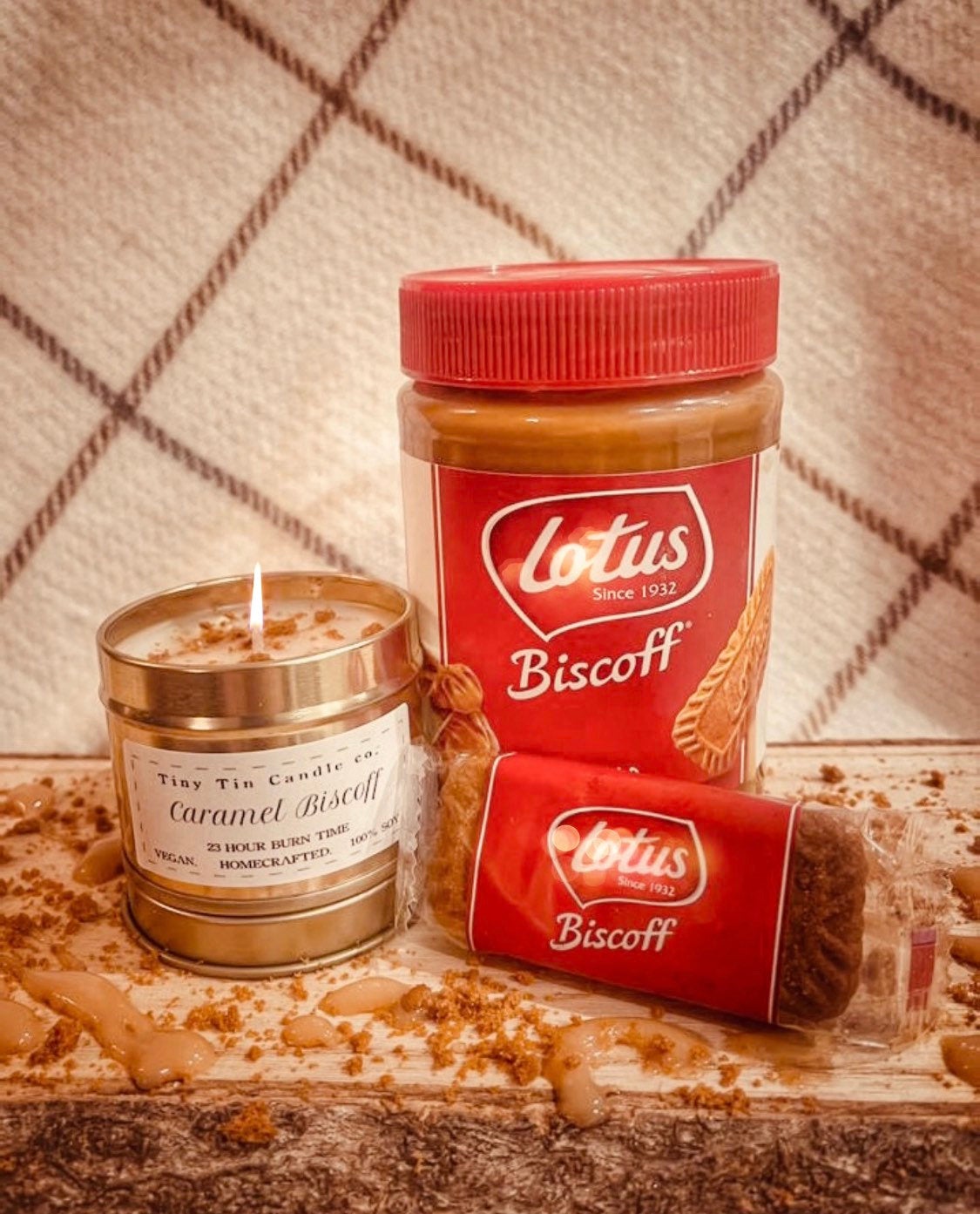 A cozy arrangement featuring a homemade candle labeled 'Caramel Biscoff' by Tiny Tin Candle with a 23-hour burn time, advertised as vegan and handcrafted. Next to it is a jar of Lotus Biscoff spread with its distinctive red lid, and a wrapped Lotus Biscoff cookie, all displayed on a wooden surface with crumbs and dollops of spread, enhancing the homey, handmade vibe. The soy candle adds a crafted touch to this warm, inviting scene