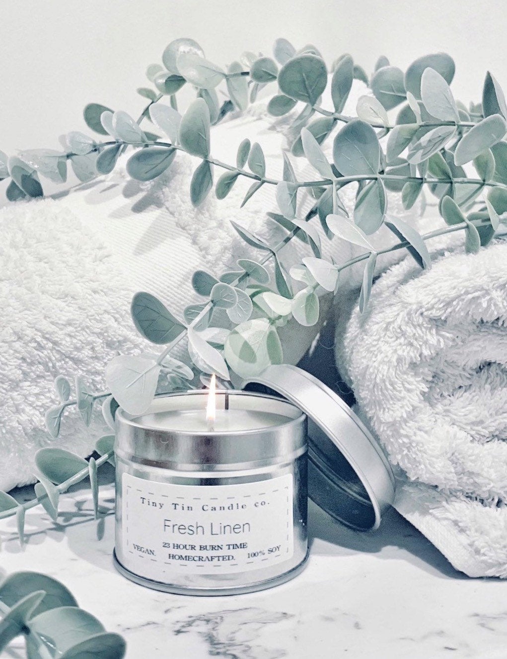 A lit homemade candle by Tiny Tin Candle Co., named Fresh Linen, with a 23-hour burn time, displayed elegantly against a backdrop of fluffy white towels and serene eucalyptus leaves. The vegan, 100% soy, handcrafted candle is encased in a sleek silver tin, exuding a sense of freshness and purity in the composition