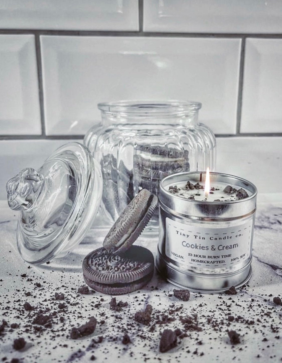 A 'Cookies & Cream' homemade soy candle from Tiny Tin Candle Co., displayed next to a glass jar of chocolate sandwich cookies, captures the sweet, comforting scent of a classic dessert treat