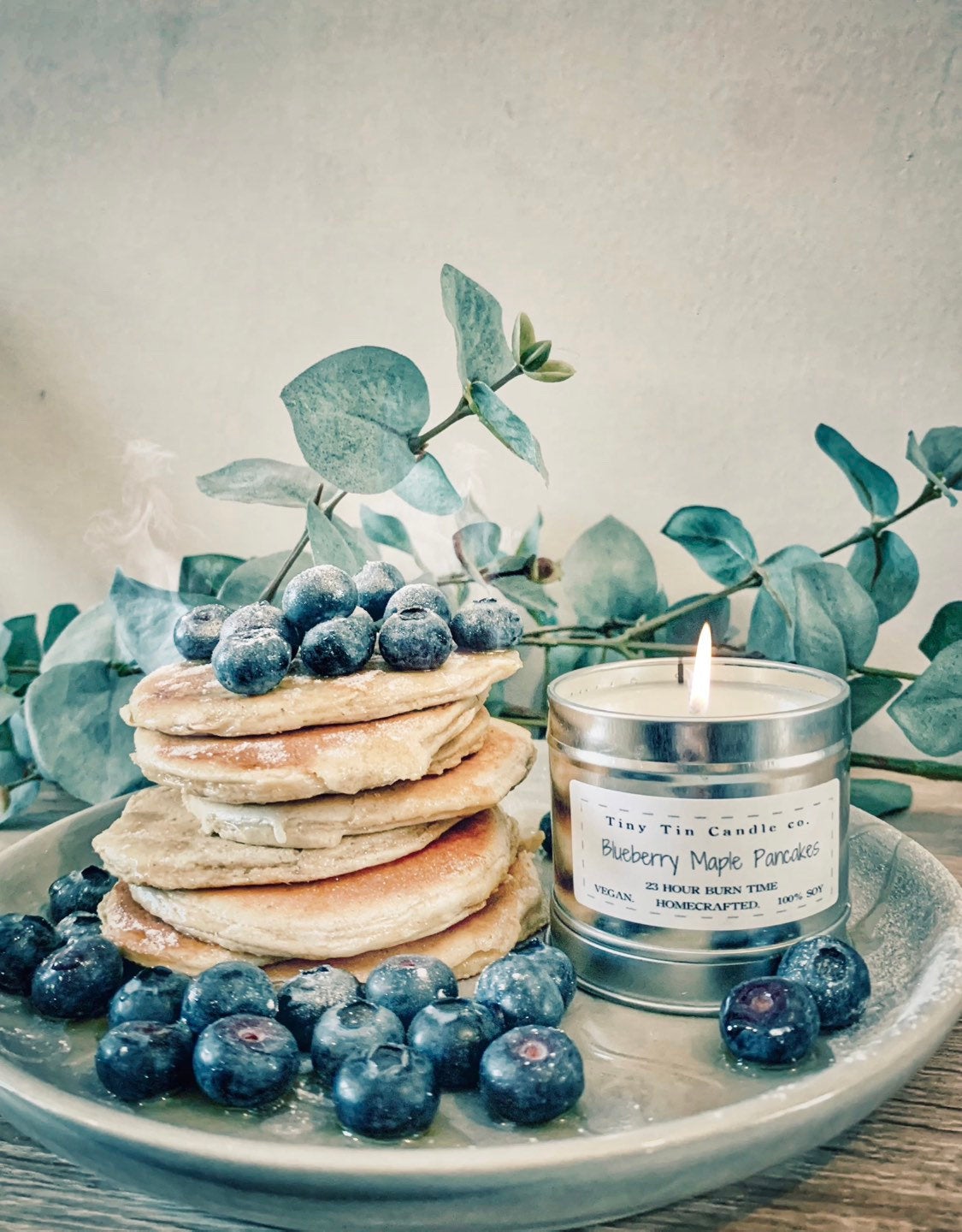 A homemade 'Blueberry Maple Pancakes' scented candle from Tiny Tin Candle Co. is lit, set against a stack of fluffy pancakes topped with fresh blueberries, embodying a homey, gourmet ambiance