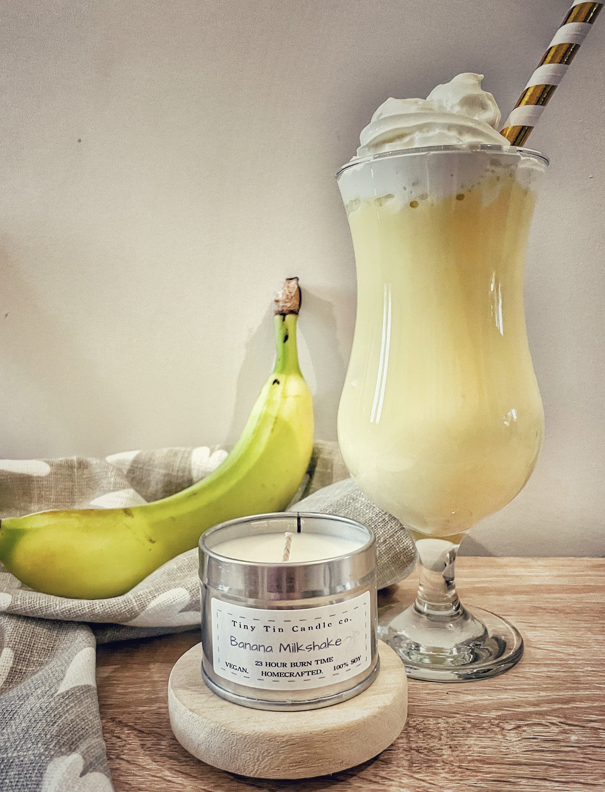 A creamy Banana Milkshake with whipped cream and a straw sits beside a ripe banana and Tiny Tin Candle Co.'s homemade 'Banana Milkshake' soy candle, lit to infuse any room with its sweet, inviting fragrance.